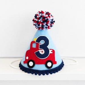May include: A light blue felt party hat featuring a red fire engine applique with a dark blue number three centred on it. The hat is topped with a multi-coloured red, white, and dark blue pom-pom and finished with a dark blue pom-pom trim.