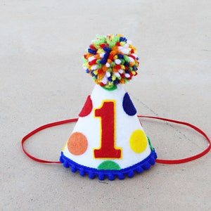 May include: A white felt party hat with a red, yellow, blue, and orange polka dot pattern. The hat has a blue pom-pom trim and a red felt chin strap. The number "1" is printed in red on the front of the hat.