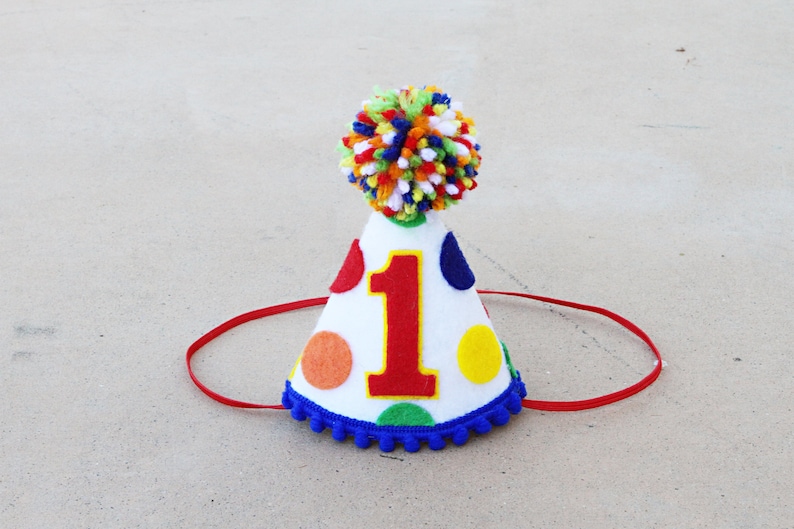 May include: A white felt party hat with a red, yellow, blue, and orange polka dot pattern. The hat has a blue pom-pom trim and a red felt chin strap. The number "1" is printed in red on the front of the hat.