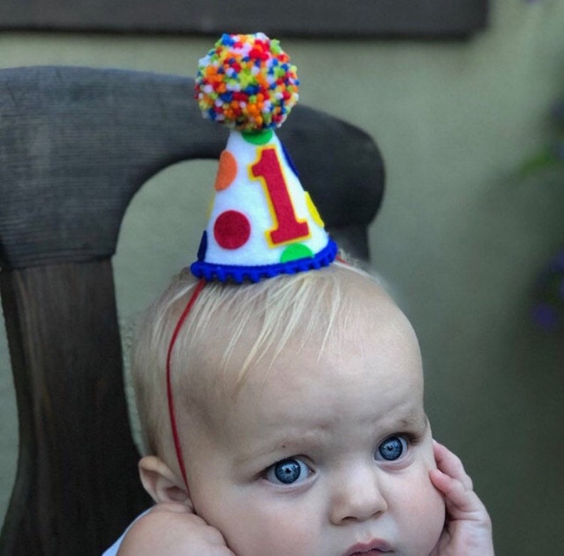 May include: A white felt party hat with a blue trim and colorful polka dots. The hat has a red felt number "1" and a colorful pom-pom on top. The hat is worn by a baby with blonde hair.