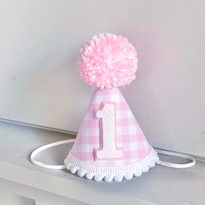 May include: A conical party hat featuring a light pink and white gingham pattern. The top is adorned with a large, fluffy pink pom-pom. A white number one is centered on the front, and the bottom edge is trimmed with a white pom-pom fringe.