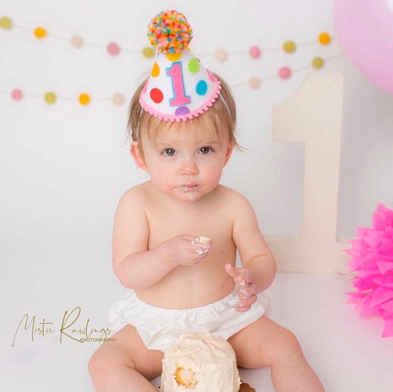 Puede incluir: Un beb&eacute; con un mono blanco y un gorro de cumplea&ntilde;os rosa y blanco con lunares de colores y el n&uacute;mero "1" en &eacute;l. El beb&eacute; est&aacute; sentado frente a un pastel blanco con glaseado blanco y una flor rosa.