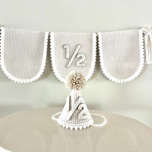 Puede incluir: Un conjunto de bander&iacute;n y gorro de fiesta para una celebraci&oacute;n de medio cumplea&ntilde;os. El bander&iacute;n tiene tres paneles festoneados con rayas verticales, ribete blanco y el texto "1/2". Tambi&eacute;n se incluye un gorro de fiesta a juego con un pomp&oacute;n y el texto "1/2".