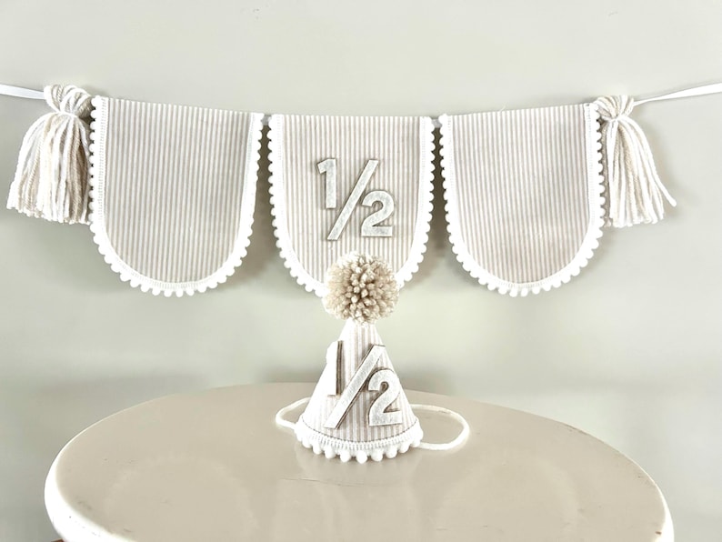 Puede incluir: Un conjunto de bander&iacute;n y gorro de fiesta para una celebraci&oacute;n de medio cumplea&ntilde;os. El bander&iacute;n tiene tres paneles festoneados con rayas verticales, ribete blanco y el texto "1/2". Tambi&eacute;n se incluye un gorro de fiesta a juego con un pomp&oacute;n y el texto "1/2".