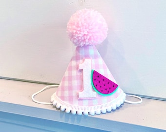 Puede incluir: Un gorro de fiesta cónico con un patrón de cuadros vichy en rosa claro y blanco, rematado con un gran pompón de lana rosa. El frente muestra un número uno de fieltro blanco y una rodaja de sandía de fieltro con centro rosa y corteza verde.