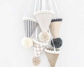 May include: A hanging mobile featuring five cone-shaped fabric ornaments suspended by white ribbons. The cones display various patterns including grey stripes, beige stripes, beige gingham, and solid tan linen, each topped with white or grey pom-poms.