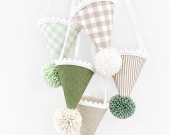 May include: A hanging mobile featuring five cone-shaped ornaments in various fabrics including beige gingham, green gingham, solid olive green, and beige stripes. Each cone is trimmed with white pom-pom fringe and a yarn pom-pom at the tip.
