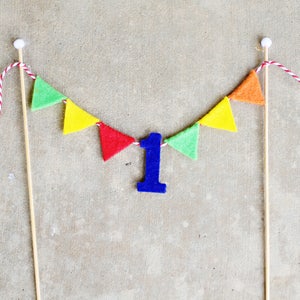 May include: A colorful felt banner with green, yellow, orange, and red triangles strung on twine. A blue felt number one is hanging in the center of the banner.