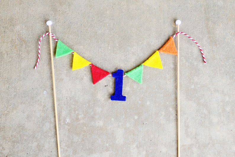 May include: A colorful felt banner with green, yellow, orange, and red triangles strung on twine. A blue felt number one is hanging in the center of the banner.