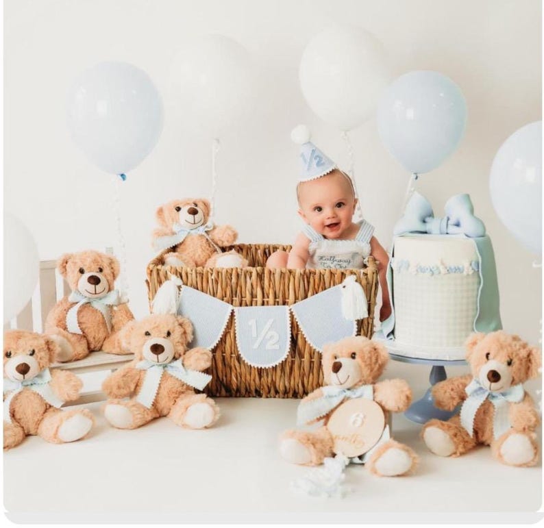 Puede incluir: Una escena de celebraci&oacute;n de medio cumplea&ntilde;os con un beb&eacute; en una cesta, osos de peluche, globos y una tarta. Las decoraciones son en azul claro y blanco. El beb&eacute; lleva un gorro de fiesta y un traje a rayas.