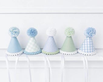 May include: Five cone-shaped party hats in soft pastel shades of blue and green. Each hat features a fluffy yarn pom-pom on top, a white pom-pom trim along the bottom edge, and long white ribbon ties. Patterns include solid, gingham, and stripes.