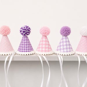 May include: Five cone-shaped party hats featuring gingham patterns in shades of pink and purple. Each hat is topped with a fluffy pom-pom and finished with a white pom-pom trim along the bottom edge, accompanied by long white ribbons for tying.