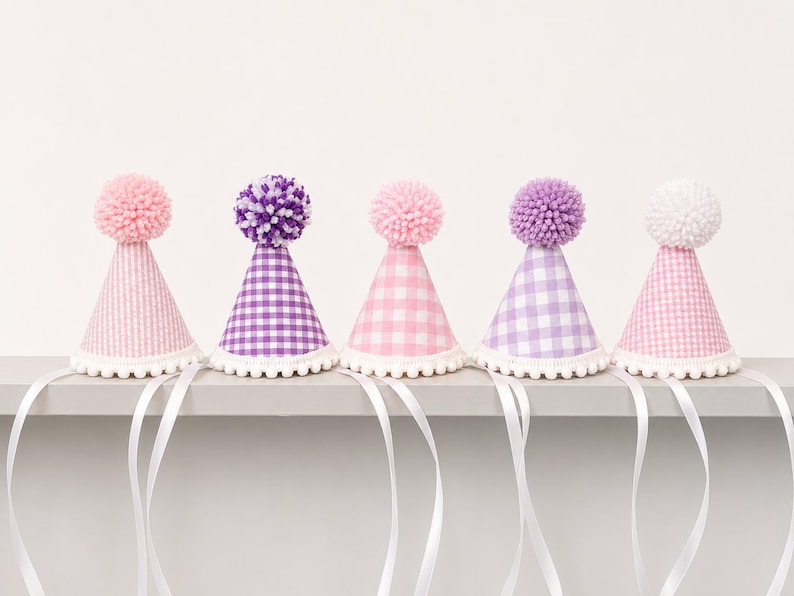 May include: Five cone-shaped party hats featuring gingham patterns in shades of pink and purple. Each hat is topped with a fluffy pom-pom and finished with a white pom-pom trim along the bottom edge, accompanied by long white ribbons for tying.