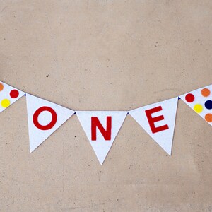 May include: A white felt banner with the word "ONE" in red letters. The banner is decorated with colorful polka dots and has blue ribbon ties on each end.