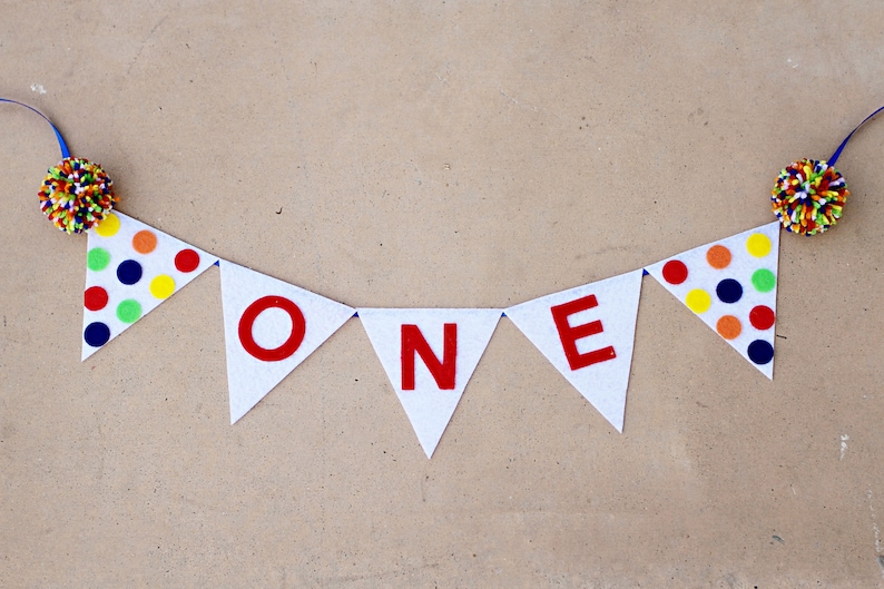 May include: A white felt banner with the word "ONE" in red letters. The banner is decorated with colorful polka dots and has blue ribbon ties on each end.