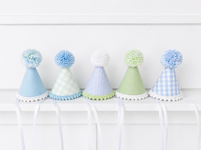 Pode incluir: Cinco chap&eacute;us de festa em forma de cone com pompons no topo e fitas brancas. Os chap&eacute;us apresentam v&aacute;rios padr&otilde;es e cores, incluindo azul-claro s&oacute;lido, verde-claro, xadrez azul e riscas verticais azuis e brancas com acabamento.