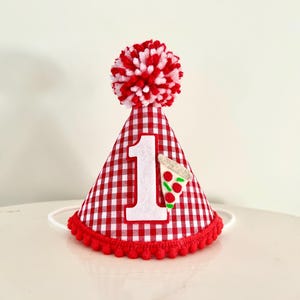 May include: Red and white gingham birthday party hat with a large white number 1 and a felt pizza slice. The hat has a red pom-pom on top and a red pom-pom trim around the base.