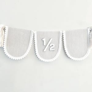 Puede incluir: Un bander&iacute;n decorativo con tres banderines con bordes festoneados. Los banderines tienen un patr&oacute;n de rayas grises y blancas y est&aacute;n unidos con una cuerda blanca. El bander&iacute;n central muestra el n&uacute;mero "1/2". Borlas blancas est&aacute;n unidas a cada extremo.