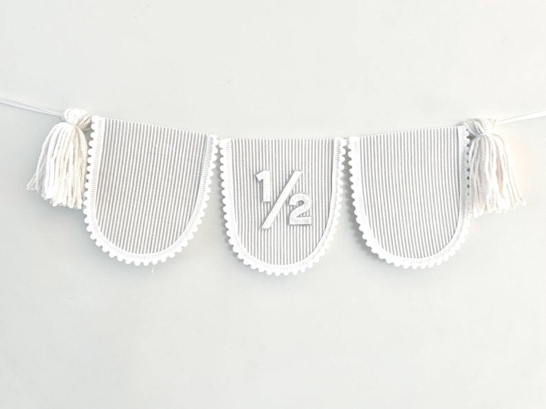 Puede incluir: Un bander&iacute;n decorativo con tres banderines con bordes festoneados. Los banderines tienen un patr&oacute;n de rayas grises y blancas y est&aacute;n unidos con una cuerda blanca. El bander&iacute;n central muestra el n&uacute;mero "1/2". Borlas blancas est&aacute;n unidas a cada extremo.