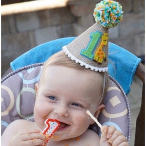 May include: A baby wearing a brown burlap party hat with a green felt number one and a yellow giraffe applique. The hat has a white pom-pom on top. The baby is holding a red and white number one candle and is smiling.