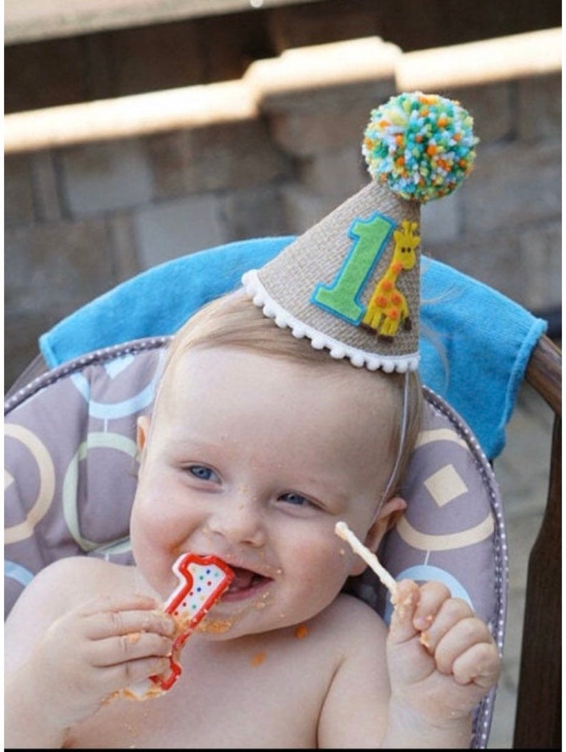 May include: A baby wearing a brown burlap party hat with a green felt number one and a yellow giraffe applique. The hat has a white pom-pom on top. The baby is holding a red and white number one candle and is smiling.