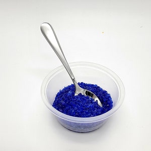 May include: A small clear plastic container filled with blue silica gel beads. A silver teaspoon is resting on top of the beads.