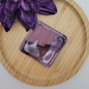 Puede incluir: Un posavasos cuadrado de vidrio púrpura con bordes redondeados se encuentra sobre una bandeja redonda de bambú marrón claro. Una gran flor de tela púrpura oscuro está colocada cerca del posavasos. El fondo es blanco.