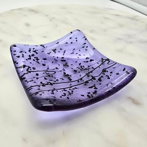 May include: A square, lavender-colored glass dish with black speckles and wavy lines. The dish has a slightly curved shape and is likely handmade. The design is simple yet elegant, suitable for holding small items.