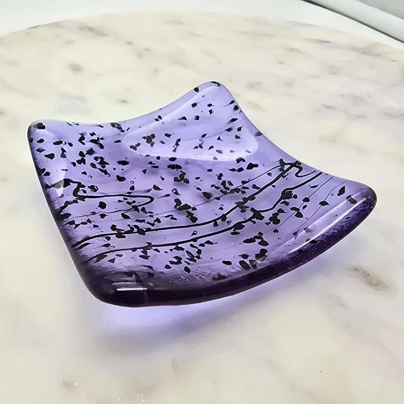 May include: A square, lavender-colored glass dish with black speckles and wavy lines. The dish has a slightly curved shape and is likely handmade. The design is simple yet elegant, suitable for holding small items.
