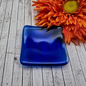 May include: A square, cobalt blue glass dish with vertical black lines. The dish has rounded corners and sits on a light wood surface. An orange flower with a yellow centre is in the background.