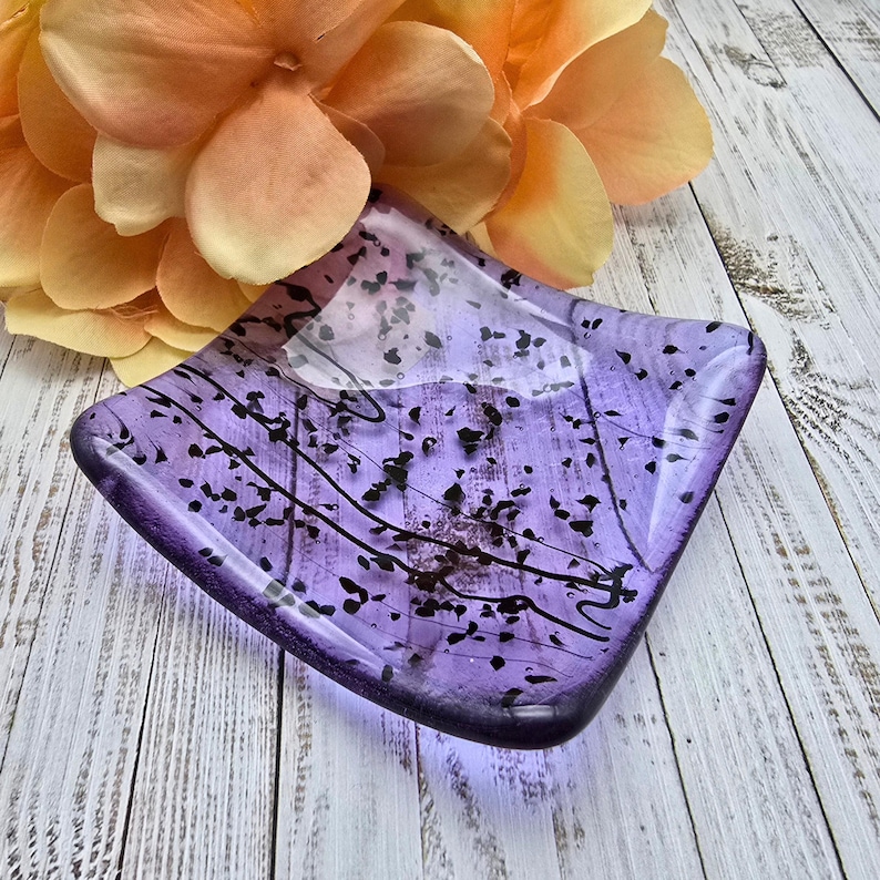 May include: A square, lavender glass dish with black speckles and lines. The dish has a slightly raised edge and sits on a light-colored wooden surface. A large, orange and yellow flower is in the background.