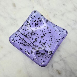 May include: A square, lavender-colored glass dish with black speckles and wavy lines. The dish has a slightly raised edge and sits on a white marble surface. The glass has a glossy finish.