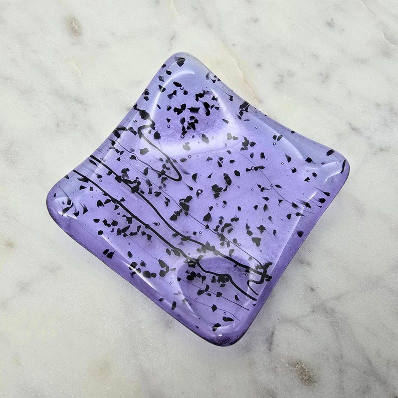 May include: A square, lavender-colored glass dish with black speckles and wavy lines. The dish has a slightly raised edge and sits on a white marble surface. The glass has a glossy finish.