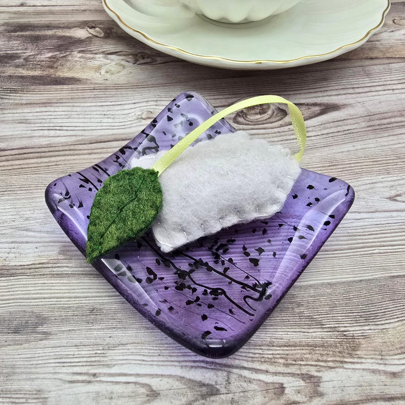 May include: A decorative, square, purple glass dish with a felt tea bag and green leaf detail. The tea bag is white with a yellow ribbon. The dish has a black, abstract design. A white teacup and saucer are in the background.