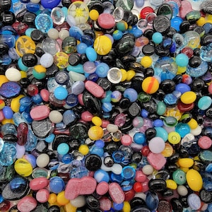 Op de afbeelding: Een grote verzameling kleurrijke glazen kiezels in verschillende vormen en maten. De kiezels zijn een mix van helder, ondoorzichtig en iriserend glas, met kleuren zoals blauw, groen, rood, geel en zwart.