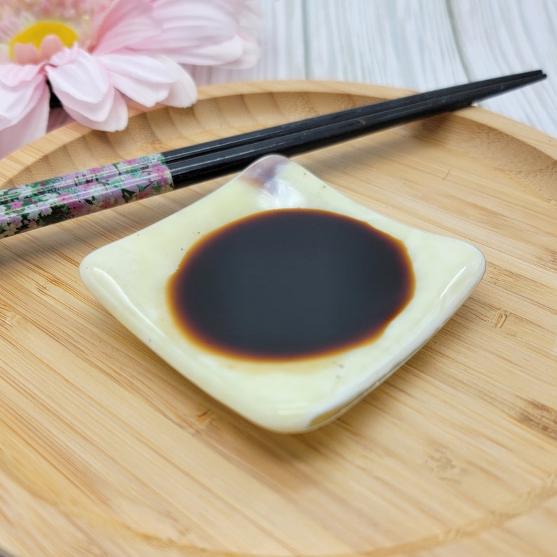 Puede incluir: Un peque&ntilde;o plato cuadrado blanco con salsa marr&oacute;n oscuro en su interior. El plato est&aacute; sobre una superficie de madera con un par de palillos con dise&ntilde;o floral.