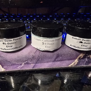Op de afbeelding: Drie kleine potjes met zwarte deksels en witte etiketten met poedervormige pigmenten. De etiketten luiden "Copper Oxide Powder", "Copper Carbonate Powder" en "Cobalt Oxide Powder". Elk potje is gelabeld met het gewicht in ounces en de Etsy shopnaam.