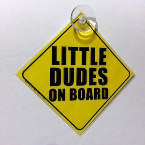 May include: Yellow diamond-shaped car sign with black text that reads "Little Dudes On Board". The sign has a suction cup on the top.