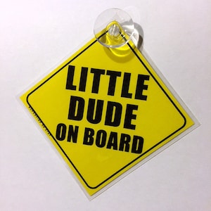 May include: Yellow diamond-shaped car sign with a suction cup. The sign reads "Little Dude On Board" in black text.
