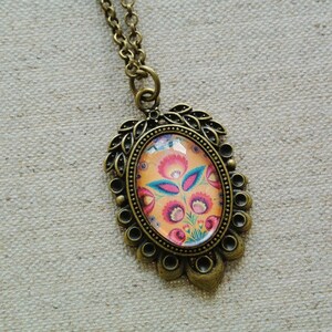 Op de afbeelding: Een gouden ketting met een ovale hanger met een kleurrijk bloemenmotief. De hanger is omgeven door een decoratieve rand van bladeren en kralen.