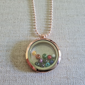 Puede incluir: Un collar con un dije de medallón de oro rosa y un inserto de vidrio transparente. El medallón contiene una variedad de cuentas de colores, incluyendo verde, azul, rojo y amarillo.