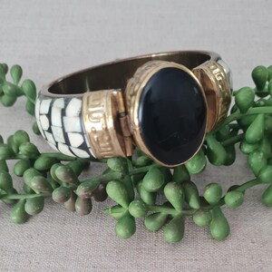 Könnte beinhalten: Ein aufklappbarer Armreif mit einem großen, zentralen ovalen schwarzen Stein in einer goldfarbenen Metallfassung. Das Armband zeigt ein geometrisches Muster aus abwechselnden weißen und schwarzen rechteckigen Kacheln, akzentuiert durch goldfarbene Metallsegmente.