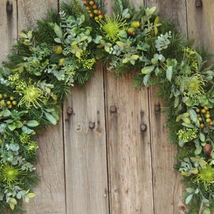 May include: A vibrant green garland featuring a mix of foliage, such as pine needles, berries, and distinctive spiky green flowers. The garland is shaped in an arch, ideal for adorning a door or wall. The backdrop is a weathered wooden door.