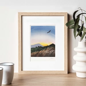 May include: A framed watercolor painting of a sunset landscape with a bird flying in the sky. The painting features a mountain range in the distance, a field of tall grass in the foreground, and a bright orange sunset.