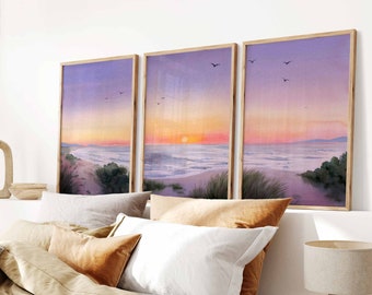 May include: Three framed watercolor paintings depicting a beach scene at sunset. The paintings are in shades of purple, pink, and orange, with a few birds flying in the sky. The beach is sandy with some green grass and bushes in the foreground.