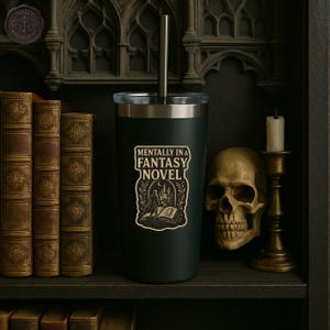 May include: A black tumbler with a silver rim and straw, featuring a sticker that reads "Mentally in a Fantasy Novel." The tumbler is displayed on a shelf with antique books, a skull, and a candle holder.