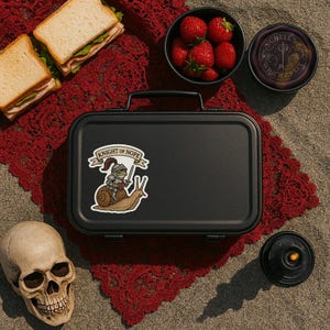 May include: A black lunchbox with a sticker that reads "Knight of Nope" featuring a cartoon knight riding a snail. The image also includes a skull, sandwiches, strawberries, and a small jar with text.