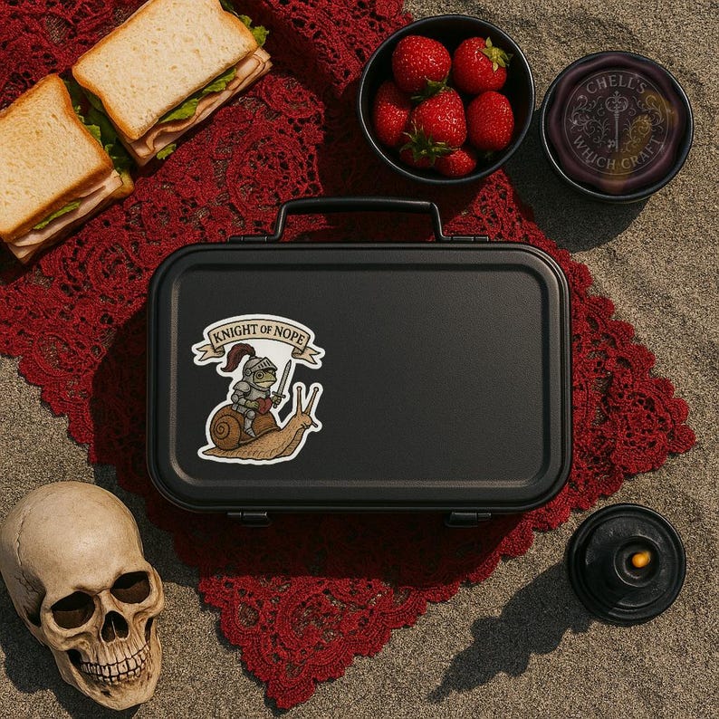 May include: A black lunchbox with a sticker that reads "Knight of Nope" featuring a cartoon knight riding a snail. The image also includes a skull, sandwiches, strawberries, and a small jar with text.