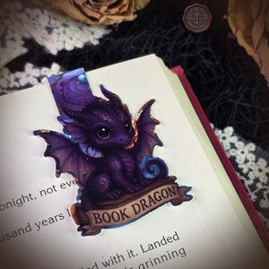 May include: A purple dragon bookmark featuring a small creature with large eyes and wings perched on a banner that reads BOOK DRAGON. The illustration is rendered in shades of deep violet and indigo with intricate patterns on the dragon's body.