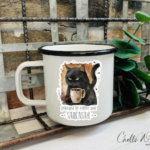 Puede incluir: Taza de esmalte blanco con borde y asa negros. La taza tiene una pegatina de un gato negro sosteniendo una taza de caf&eacute;, con el texto "powered by coffee and sarcasm". Un art&iacute;culo humor&iacute;stico.